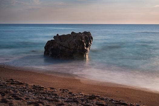 View of a large dark rock formation stands defiant against the soft blurred turquoise sea under a pastel sky on a sandy beach Lefkada Greece. View of a large dark rock formation stands defiant against the soft blurred turquoise sea under a pastel sky on a sandy beach Lefkada Greece.
