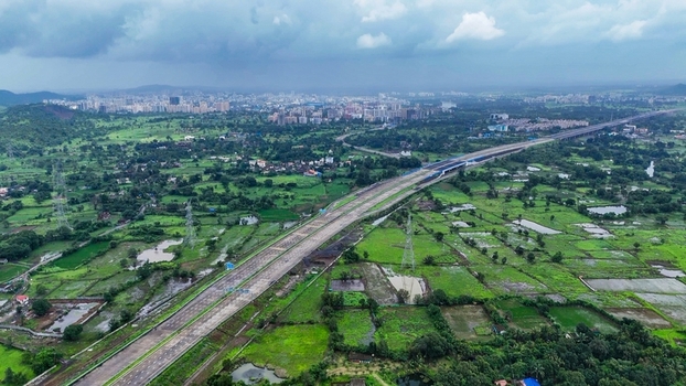 Aerial view of the Delhi Mumbai Expressway cutting through lush green fields and rural landscape with a distant city skyline under a cloudy sky Badlapur Maharashtra India. Aerial view of the Delhi Mumbai Expressway cutting through lush green fields and rural landscape with a distant city skyline under a cloudy sky Badlapur Maharashtra India.