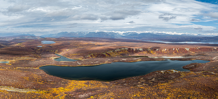 Aerial view of the tundra landscape near Paxson featuring dark blue lakes autumn vegetation and snow capped mountains under a cloudy sky in Paxson Alaska United States.