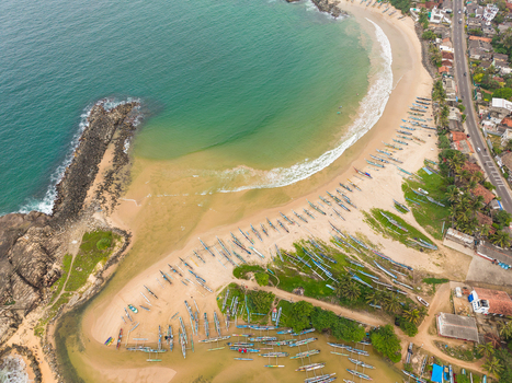 Aerial view of traditional outrigger fishing boats lined up on the sandy beach of a tropical bay with turquoise water and a rocky breakwater Dodanduwa Southern Province Sri Lanka. Aerial view of traditional outrigger fishing boats lined up on the sandy beach of a tropical bay with turquoise water and a rocky breakwater Dodanduwa Southern Province Sri Lanka.