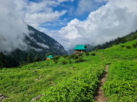 View of a traditional house with a green roof nestled in the lush green mountains of Malana surrounded by rolling hills a dirt path and dramatic clouds Malana Himachal Pradesh India.