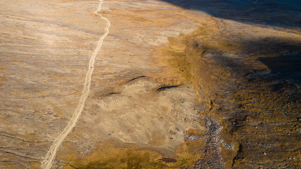 Aerial view of the vast Deosai Plains with a winding dirt road cutting through the golden landscape under dramatic shadows in Skardu Gilgit Pakistan.