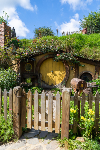 View of a quaint hobbit hole with a vibrant yellow door nestled into a grassy hill framed by a rustic wooden fence Hobbiton Movie Set Waikato Region New Zealand. View of a quaint hobbit hole with a vibrant yellow door nestled into a grassy hill framed by a rustic wooden fence Hobbiton Movie Set Waikato Region New Zealand.