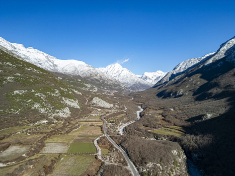 Aerial view of the mountain valley featuring snow capped peaks winding roads and agricultural fields under a clear blue sky in Bzhete Markaj Qarku i Shkodres Albania. Aerial view of the mountain valley featuring snow capped peaks winding roads and agricultural fields under a clear blue sky in Bzhete Markaj Qarku i Shkodres Albania.