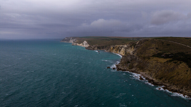 Aerial view of Lulworth Cove featuring dramatic limestone cliffs turquoise waters and rugged coastline under a cloudy sky in the United Kingdom.