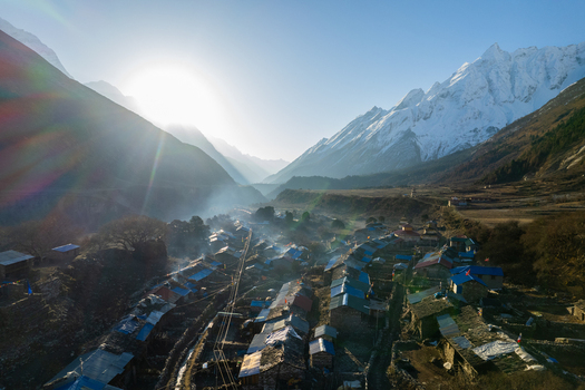 Aerial view of a remote village nestled in a Himalayan valley with snow capped peaks rising smoke from stone houses and bright sunlight in Nepal.