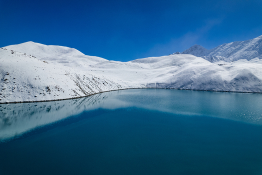 Aerial view of a pristine alpine lake surrounded by snow covered mountain peaks under a clear blue sky in Nepal.