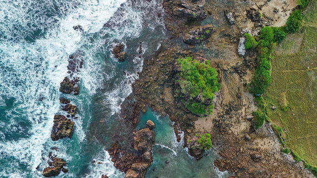 Aerial view of a rocky coastline with turquoise waves crashing against rugged cliffs and lush green vegetation along the shore in Indonesia. Aerial view of a rocky coastline with turquoise waves crashing against rugged cliffs and lush green vegetation along the shore in Indonesia.