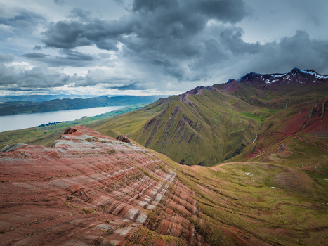 Aerial view of the striated rock formations of Pallay Punchu Rainbow Mountain with green slopes and Langui Layo lake under a dramatic cloudy sky in Layo Cuzco Peru.