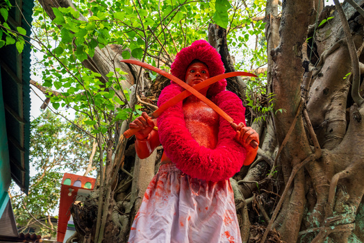 Munshiganj Bangladesh 13 April 2026 View of a man covered in red paint holding crossed swords and wearing a large pink garland during a traditional festival in front of a banyan tree.