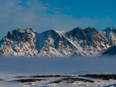 Aerial view of snow covered jagged mountain peaks rising above a thick layer of low lying mist and a frozen arctic landscape in Tasiilaq Sermersooq Municipality Greenland. Aerial view of snow covered jagged mountain peaks rising above a thick layer of low lying mist and a frozen arctic landscape in Tasiilaq Sermersooq Municipality Greenland.