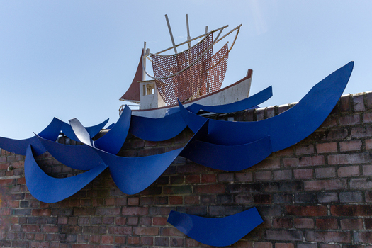 View of a nautical sculpture featuring a boat on blue metal waves mounted on a brick wall under a clear sky in Hooksiel Wangerland Niedersachsen Germany. View of a nautical sculpture featuring a boat on blue metal waves mounted on a brick wall under a clear sky in Hooksiel Wangerland Niedersachsen Germany.
