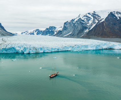 Aerial view of a sailboat navigating the turquoise waters of a glacial bay with a massive glacier wall and snow capped mountains in the background in Greenland.