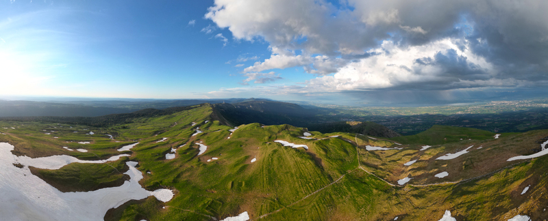 Aerial view of Le Colomby de Gex featuring green mountain ridges with patches of melting snow under a dramatic cloudy sky in Echenevex Auvergne Rhone Alpes France. Aerial view of Le Colomby de Gex featuring green mountain ridges with patches of melting snow under a dramatic cloudy sky in Echenevex Auvergne Rhone Alpes France.