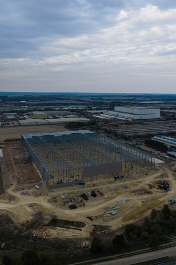 Aerial view of a large industrial construction site featuring a steel frame structure surrounding warehouses and dirt ground under a cloudy sky in United Kingdom. Aerial view of a large industrial construction site featuring a steel frame structure surrounding warehouses and dirt ground under a cloudy sky in United Kingdom.