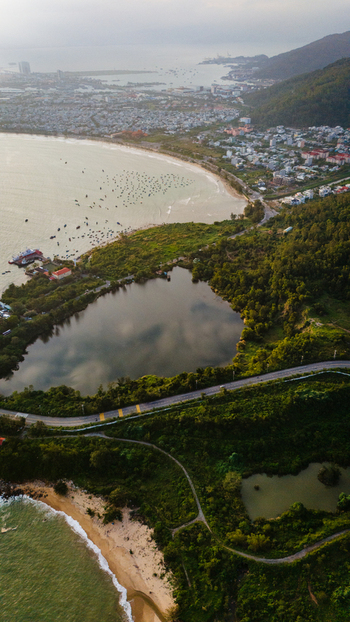 Aerial view of the coastal landscape featuring a calm lake lush green hills a winding coastal road and sandy beach meeting the sea with small boats in Da Nang Vietnam. Aerial view of the coastal landscape featuring a calm lake lush green hills a winding coastal road and sandy beach meeting the sea with small boats in Da Nang Vietnam.