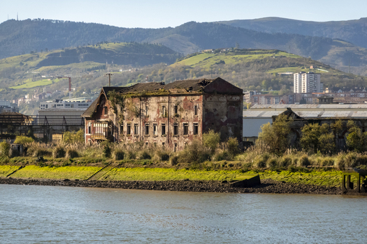 View of an abandoned industrial building with a collapsed roof and decaying walls on the riverbank against rolling green hills Greater Bilbao Basque Country Spain. View of an abandoned industrial building with a collapsed roof and decaying walls on the riverbank against rolling green hills Greater Bilbao Basque Country Spain.