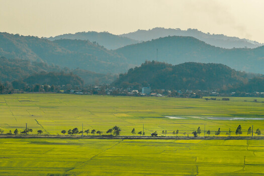 Aerial view of Lak Lake surrounded by vibrant green rice fields a line of trees along a rural road and rolling mountains in the distance in Lien Son Lak Dak Lak Vietnam. Aerial view of Lak Lake surrounded by vibrant green rice fields a line of trees along a rural road and rolling mountains in the distance in Lien Son Lak Dak Lak Vietnam.