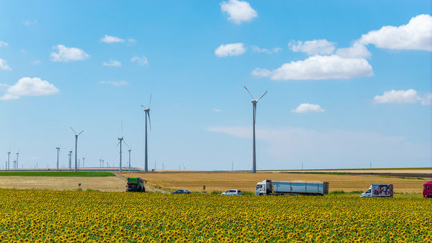 Aerial view of wind turbines rising above a vibrant sunflower field with trucks driving along a road in the countryside of Tulcea Tulcea Romania.