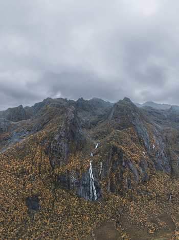 Aerial view of the dramatic Lofoten mountains featuring a cascading waterfall amidst vibrant autumn foliage under a cloudy sky in Leknes Nordland Norway. Aerial view of the dramatic Lofoten mountains featuring a cascading waterfall amidst vibrant autumn foliage under a cloudy sky in Leknes Nordland Norway.