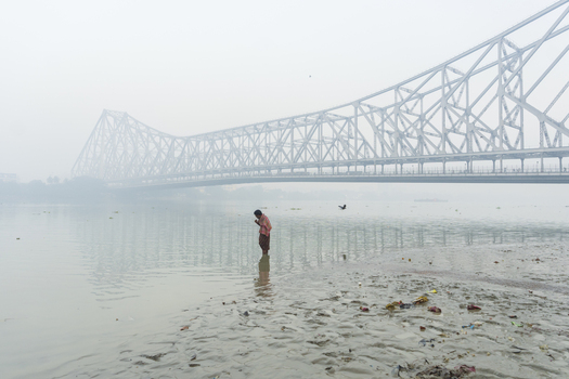 View of a solitary figure stands waist deep in the tranquil river their hands clasped in prayer beneath the colossal Howrah Bridge veiled in mist Kolkata West Bengal India. View of a solitary figure stands waist deep in the tranquil river their hands clasped in prayer beneath the colossal Howrah Bridge veiled in mist Kolkata West Bengal India.