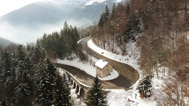 Aerial view of Col de la Faucille showing a winding mountain road with a small building on a sharp curve surrounded by snow covered pine forests in Gex Auvergne Rhone Alpes France. Aerial view of Col de la Faucille showing a winding mountain road with a small building on a sharp curve surrounded by snow covered pine forests in Gex Auvergne Rhone Alpes France.