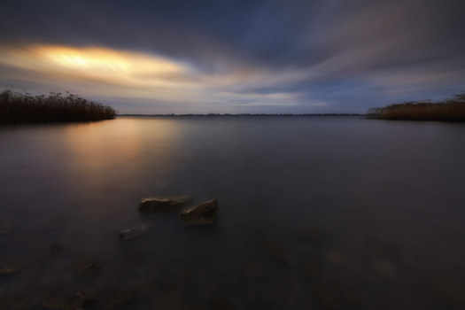 View of Lake Tarnobrzeg with calm water foreground rocks and reeds under a dramatic sunset sky in Tarnobrzeg Podkarpackie Voivodeship Poland. View of Lake Tarnobrzeg with calm water foreground rocks and reeds under a dramatic sunset sky in Tarnobrzeg Podkarpackie Voivodeship Poland.