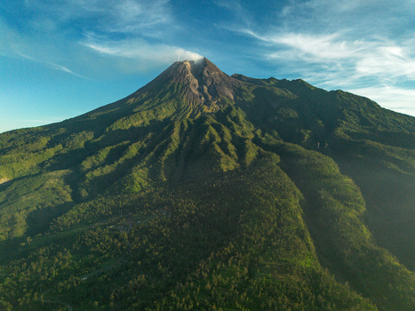 Aerial view of Mount Merapi with lush green slopes and volcanic crater under a clear blue sky in Indonesia.