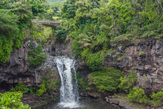 View of the Upper Waikani Falls cascading down rocky cliffs surrounded by lush tropical vegetation and a stone bridge in the background in Maui County Hawaii United States.