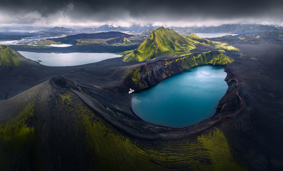 Aerial view of a turquoise volcanic crater lake surrounded by mossy green mountains and dark volcanic soil under dramatic clouds in Iceland.