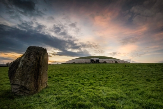 View of the prehistoric Newgrange passage tomb with a large standing stone in the foreground under a dramatic sunset sky in Donore County Meath Ireland.