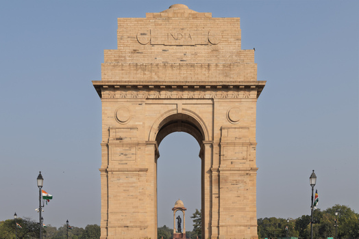 View of the majestic India Gate stands tall carved with INDIA on top framed by a clear blue sky and distant architecture visible through the arch Delhi India. View of the majestic India Gate stands tall carved with INDIA on top framed by a clear blue sky and distant architecture visible through the arch Delhi India.