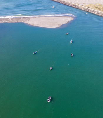 Aerial view of salmon fishing boats navigating the blue green waters of the Rogue River at a sandbar and jetty in Gold Beach Oregon United States.