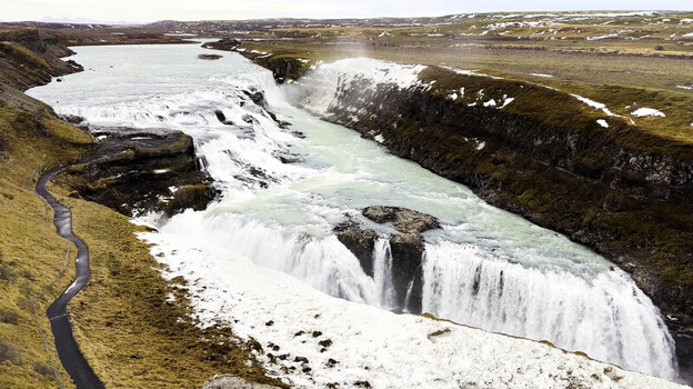 Aerial view of the powerful Gullfoss waterfall cascading through a rocky canyon with snow dusted terrain and a winding path in Blaskogabyggo Iceland.