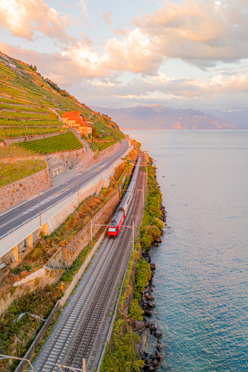 Aerial view of a train traveling along tracks beside Lake Geneva with terraced vineyards a coastal road and mountains under a warm sunset sky in Bourg en Lavaux Vaud Switzerland. Aerial view of a train traveling along tracks beside Lake Geneva with terraced vineyards a coastal road and mountains under a warm sunset sky in Bourg en Lavaux Vaud Switzerland.