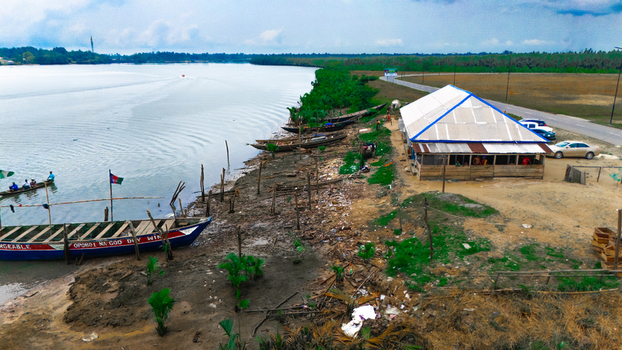 Aerial view of wooden boats moored along the muddy riverbank near a small building with a white roof a road with parked cars and lush vegetation in Minima Rivers Nigeria.