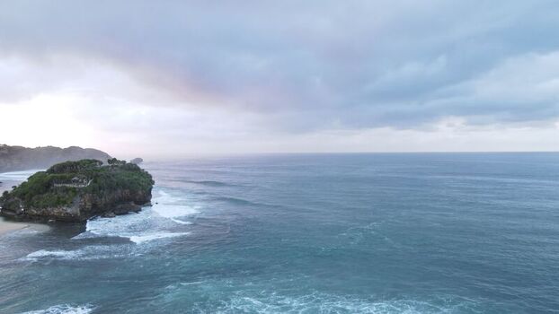 Aerial view of Pantai Drini a rocky islet with lush vegetation surrounded by the Indian Ocean under a cloudy sky in Kecamatan Tanjungsari Daerah Istimewa Yogya Indonesia. Aerial view of Pantai Drini a rocky islet with lush vegetation surrounded by the Indian Ocean under a cloudy sky in Kecamatan Tanjungsari Daerah Istimewa Yogya Indonesia.