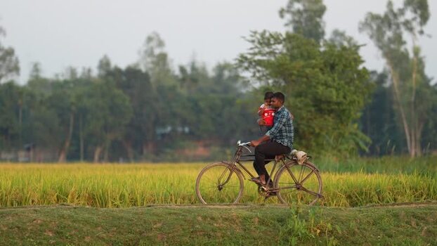 View of a man riding a bicycle with a child through fields Bangladesh.