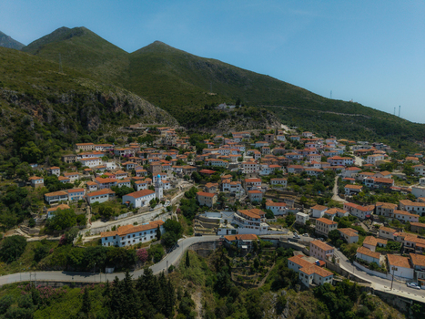 Aerial view of the hillside village featuring white houses with red roofs and a blue domed church nestled against lush green mountains in Dhermi Vlore County Albania. Aerial view of the hillside village featuring white houses with red roofs and a blue domed church nestled against lush green mountains in Dhermi Vlore County Albania.