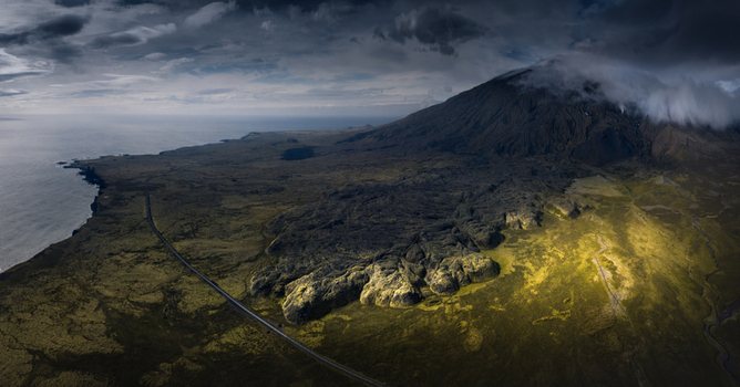 Aerial view of a volcanic mountain with clouds on its peak and vast mossy lava fields along the coastline in Iceland.