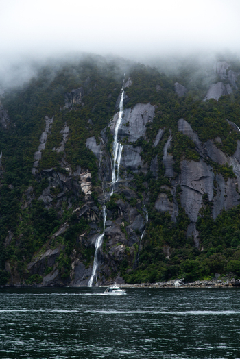 View of cascading waterfalls plummeting down rugged moss covered cliffs into the dark waters below with a boat sailing by shrouded in mist Milford Sound Southland Region New Zealand.