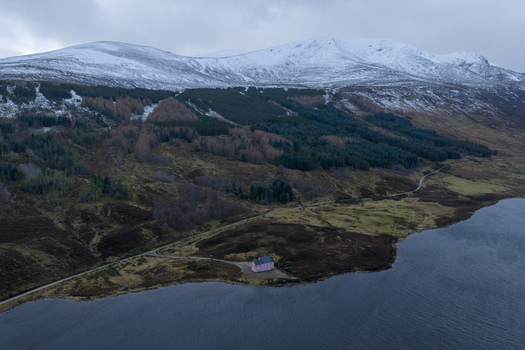 Aerial view of The Pink House on the shore of a lake with snow capped mountains and a dense forest in Dingwall Scotland United Kingdom. Aerial view of The Pink House on the shore of a lake with snow capped mountains and a dense forest in Dingwall Scotland United Kingdom.