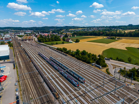 Aerial view of Daniken train station with passenger trains on multiple tracks surrounded by green fields rural landscape and blue sky with clouds in Daniken Solothurn Switzerland.