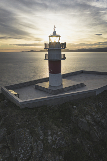 Aerial view of a lighthouse on a rocky cliff overlooking the ocean during a golden sunset with soft light reflecting on the water Galicia Spain.