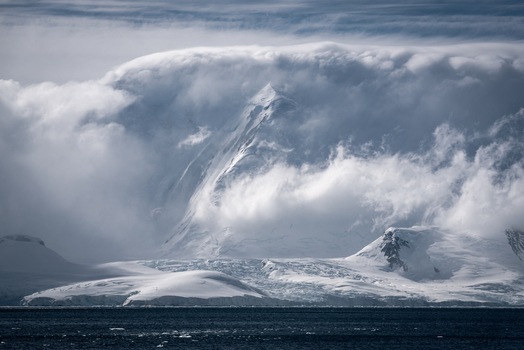 View of snow covered mountain peaks partially obscured by thick white clouds above the dark blue sea Antarctica.