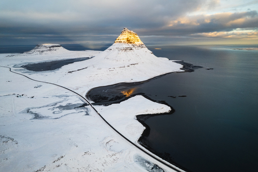 Aerial view of snow covered Kirkjufell mountain catching the golden sunlight near the dark icy waters of the coast Kirkjufell Iceland.