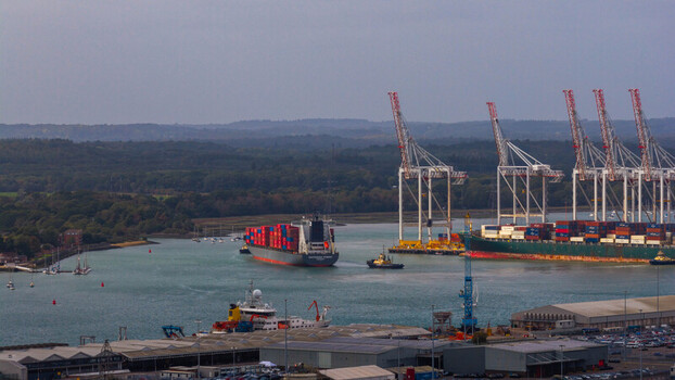 Aerial view of the Port of Southampton with a large container ship navigating the channel towering gantry cranes and cargo containers in Southampton England United Kingdom. Aerial view of the Port of Southampton with a large container ship navigating the channel towering gantry cranes and cargo containers in Southampton England United Kingdom.