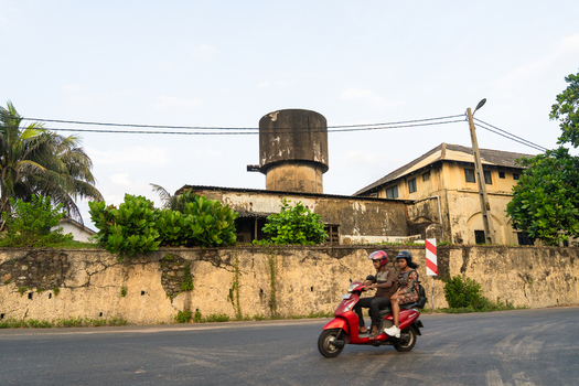 Sri Lanka 15 January 2026 View of a man and a woman riding a red scooter past a weathered concrete water tower and historic stone walls.