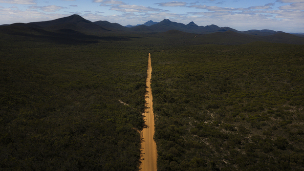 Aerial view of a long straight dirt road cutting through dense green scrubland towards distant mountains in Perth Western Australia Australia.