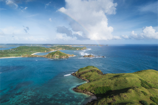 Aerial view of Tanjung Aan Beach featuring green hills and turquoise ocean water under a cloudy sky with a faint rainbow in Kecamatan Pujut Nusa Tenggara Barat Indonesia.